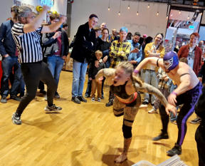 En kvinnlig wrestler blir kastad mot kameran av en manlig wrestler med ansiktet dolt av en huva. De är omgivna av folk som tittar på